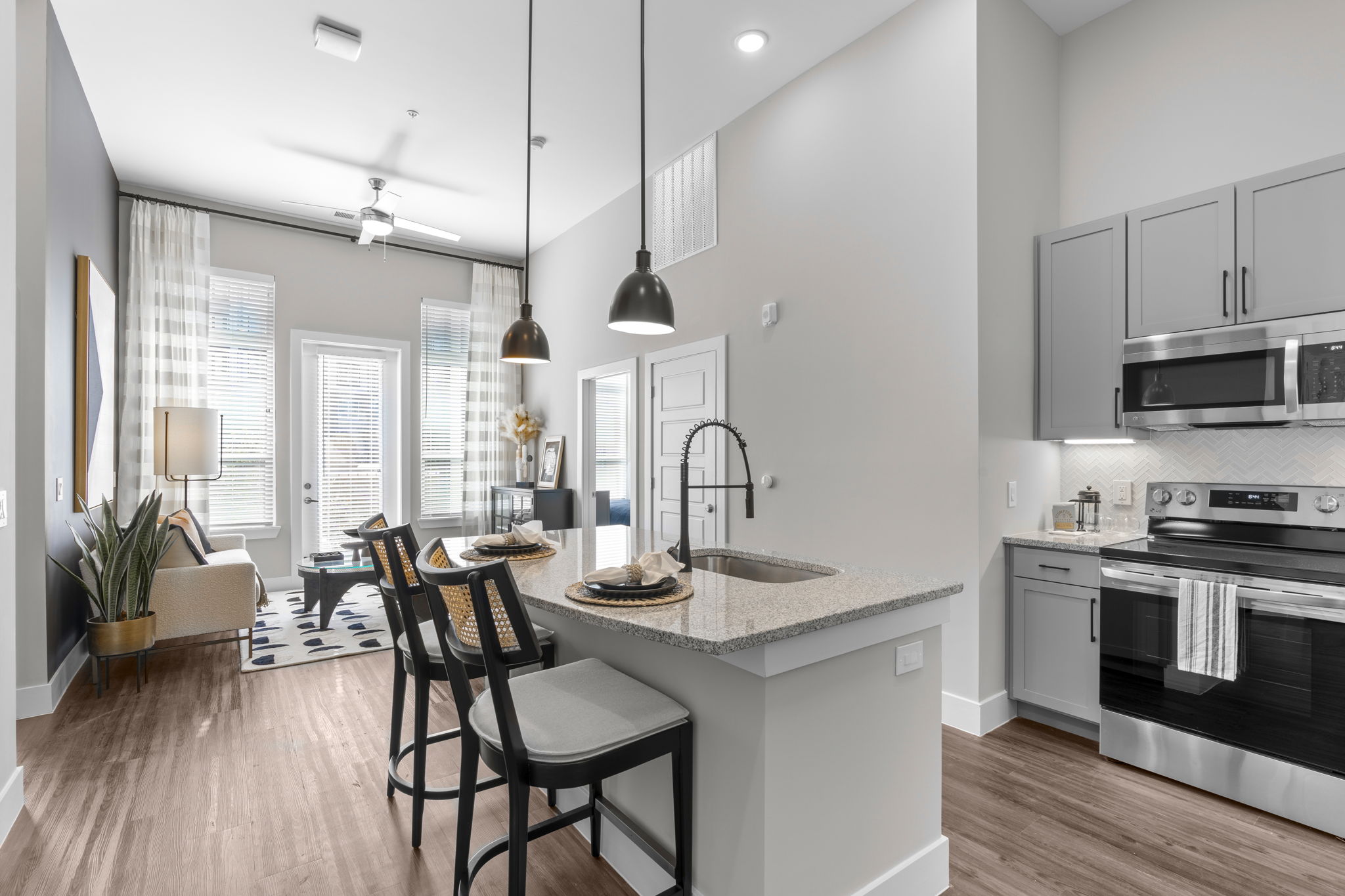 Open kitchen with gray cabinets, granite island seating, and view into living room with plants and windows